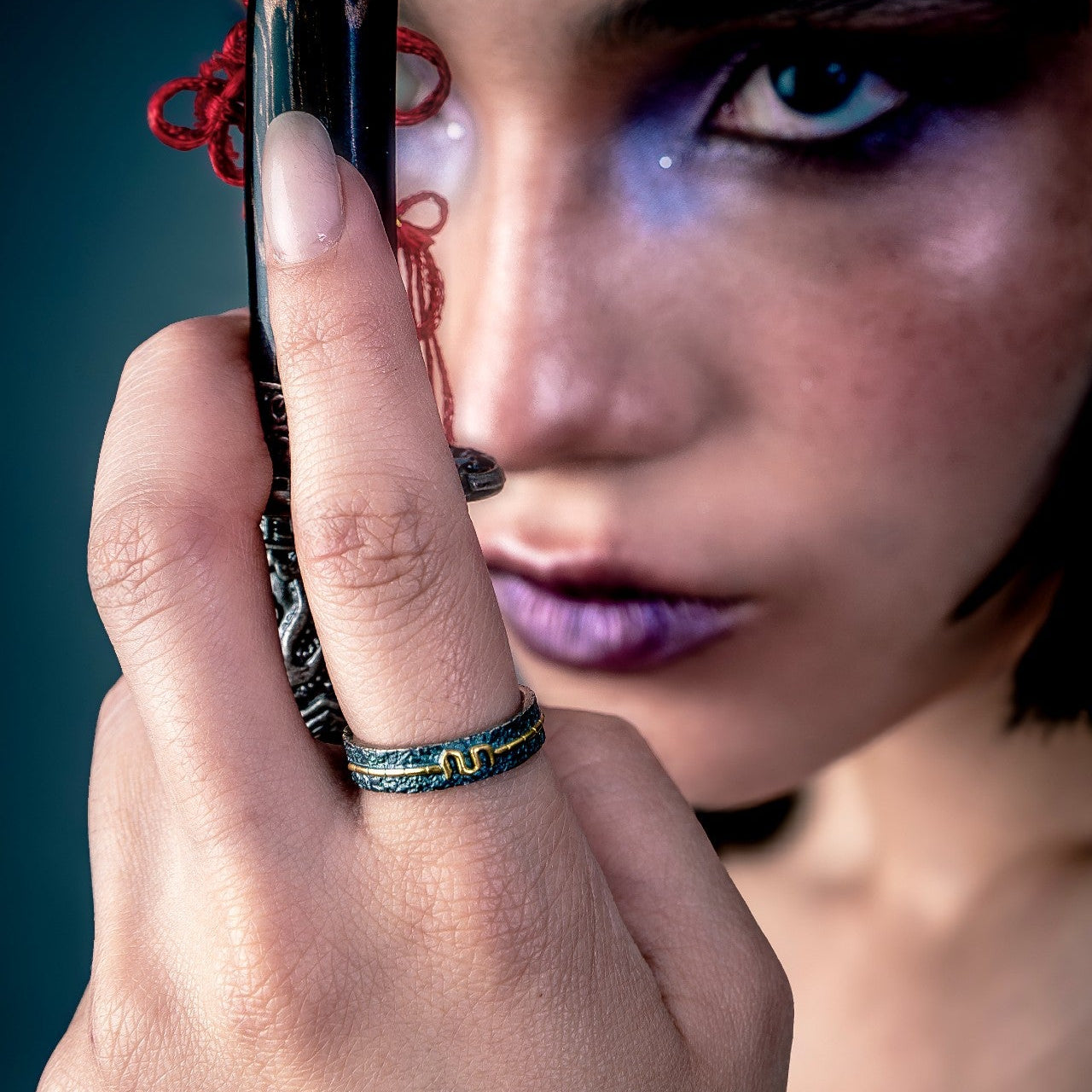 [full] Close-up of a hand wearing a statement silver ring holding a decorative object near a person's face against a dark background