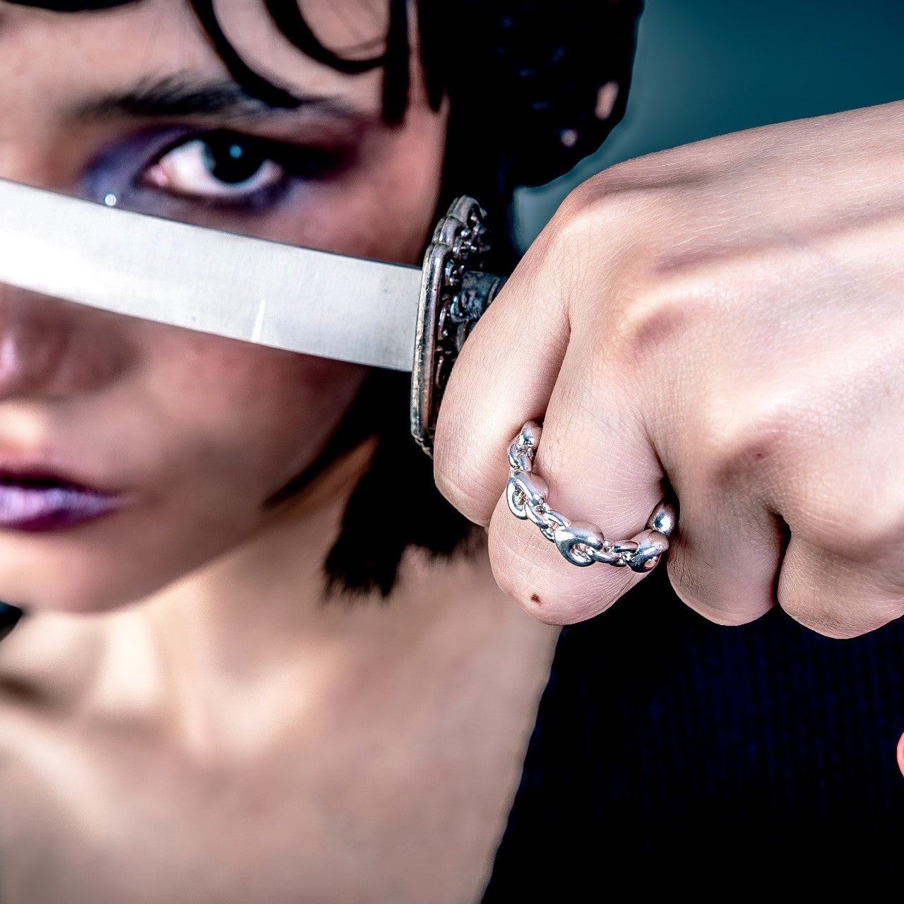 [full] Person wearing a silver ring holding a sword with a red tassel against a dark background