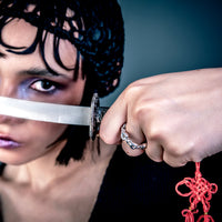 [full] Person wearing a silver ring holding a sword with a red tassel against a dark background