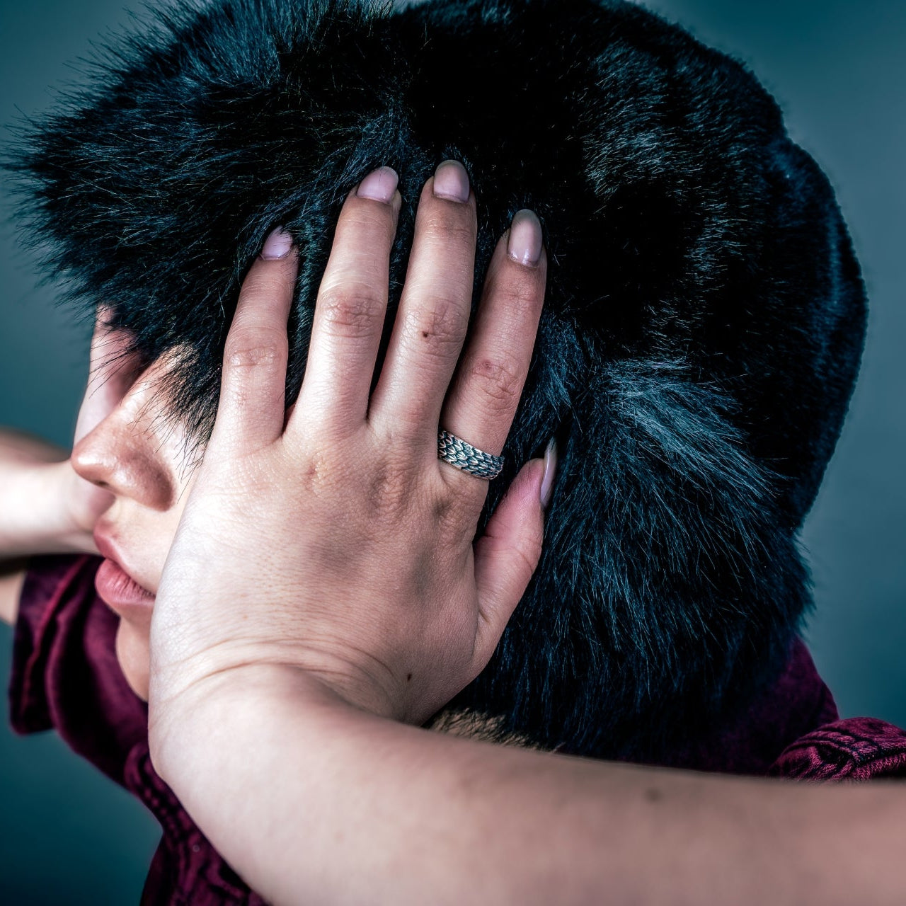 [full] - Person with a dark fur coat wearing a silver ring, covering their face against a dark background