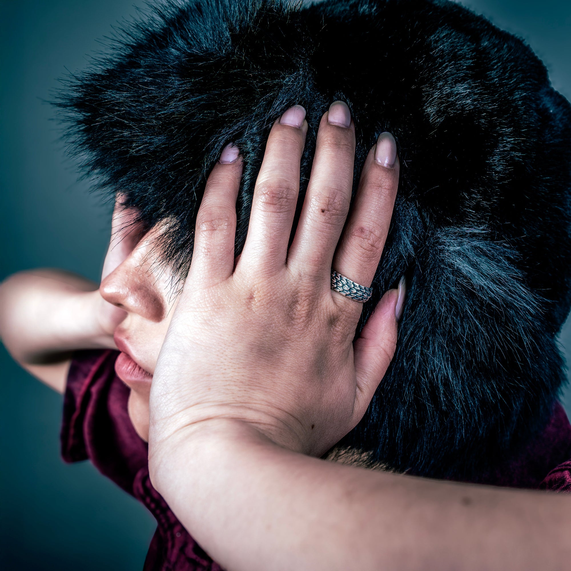 [full] - Person with a dark fur coat wearing a silver ring, covering their face against a dark background