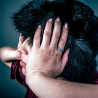 [full] - Person with a dark fur coat wearing a silver ring, covering their face against a dark background