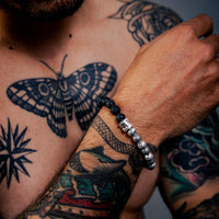 [full] Person with tattoos on chest and arms wearing black lava stone and sterling silver bead bracelet with contrasting textures.