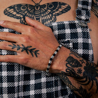 [full] - Person wearing checkered overalls with tattoos and a sterling silver star bead bracelet with black cord and polished finish.