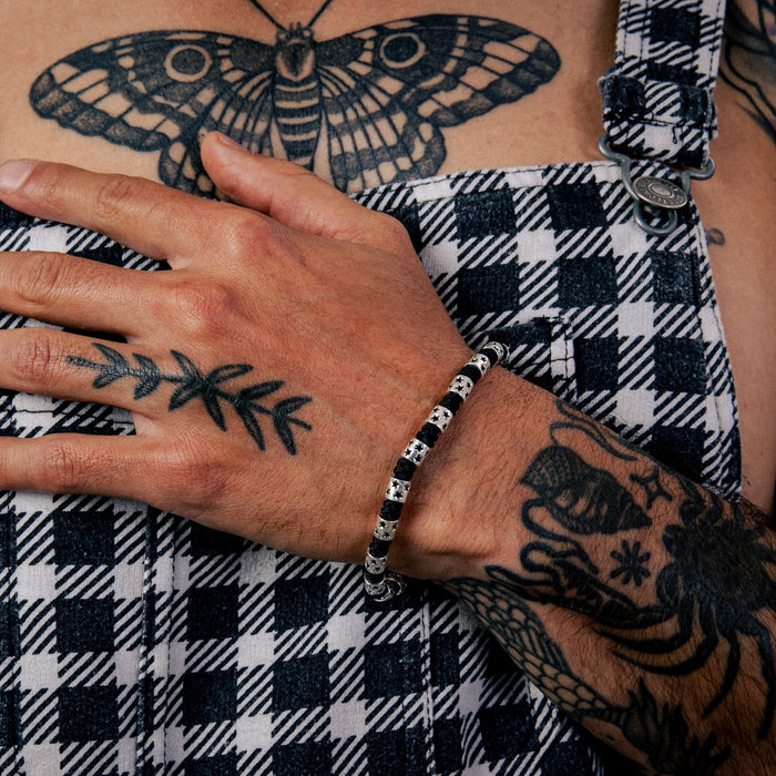 [full] - Person wearing checkered overalls with tattoos and a sterling silver star bead bracelet with black cord and polished finish.