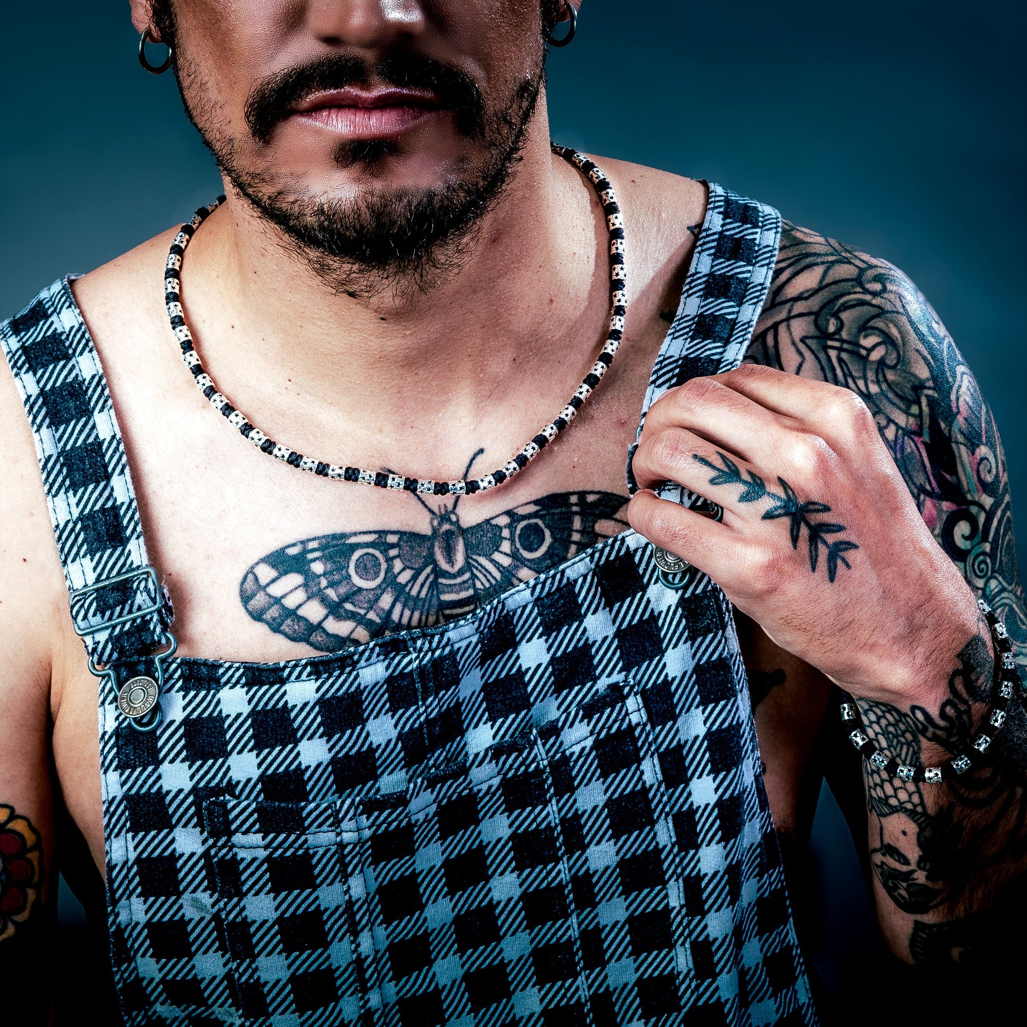 [full]- Man with tattoos wearing sterling silver and cordura necklace, and plaid overalls against a dark background