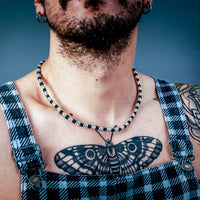 [full]- Man with tattoos wearing sterling silver and cordura necklace, and plaid overalls against a dark background