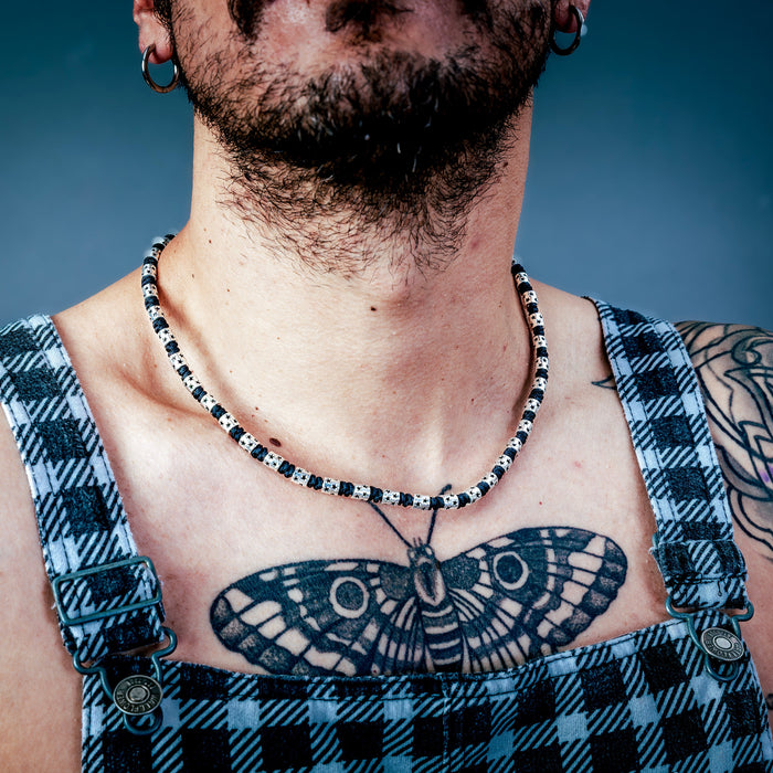 [full]- Man with tattoos wearing sterling silver and cordura necklace, and plaid overalls against a dark background