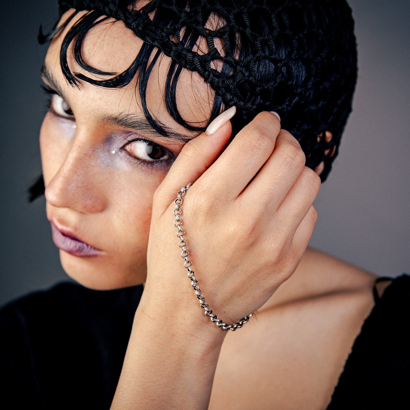 [full] Person wearing a black headpiece with decorative elements, holding their hand near their face, wearing a silver chain bracelet with a unique clasp.