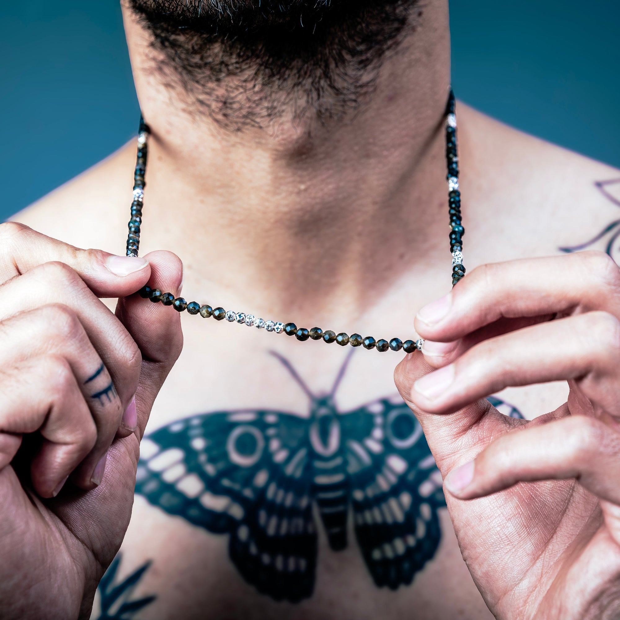 [full]-Man with a butterfly tattoo on his chest holding a beaded sterling silver necklace against a blue background