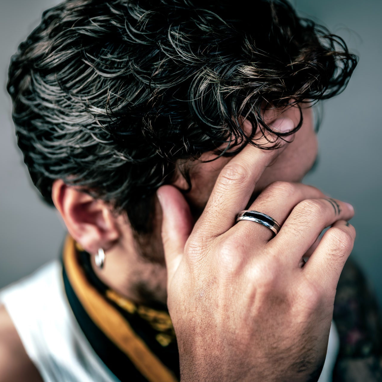 [full] Person with curly hair covering face with hand, wearing a ceramic ring, against a blurred background