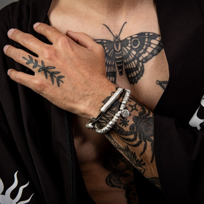 [full] Person with tattoos on arm and hand wearing a pure 999 fine silver bracelet, and a black outfit with white designs.