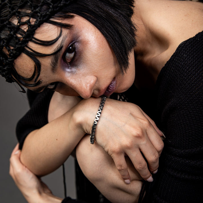 [full] - Person wearing a black outfit with thin straps and an industrial-style sterling silver bracelet with ruthenium plating and brushed gun-metal finish on a dark background.