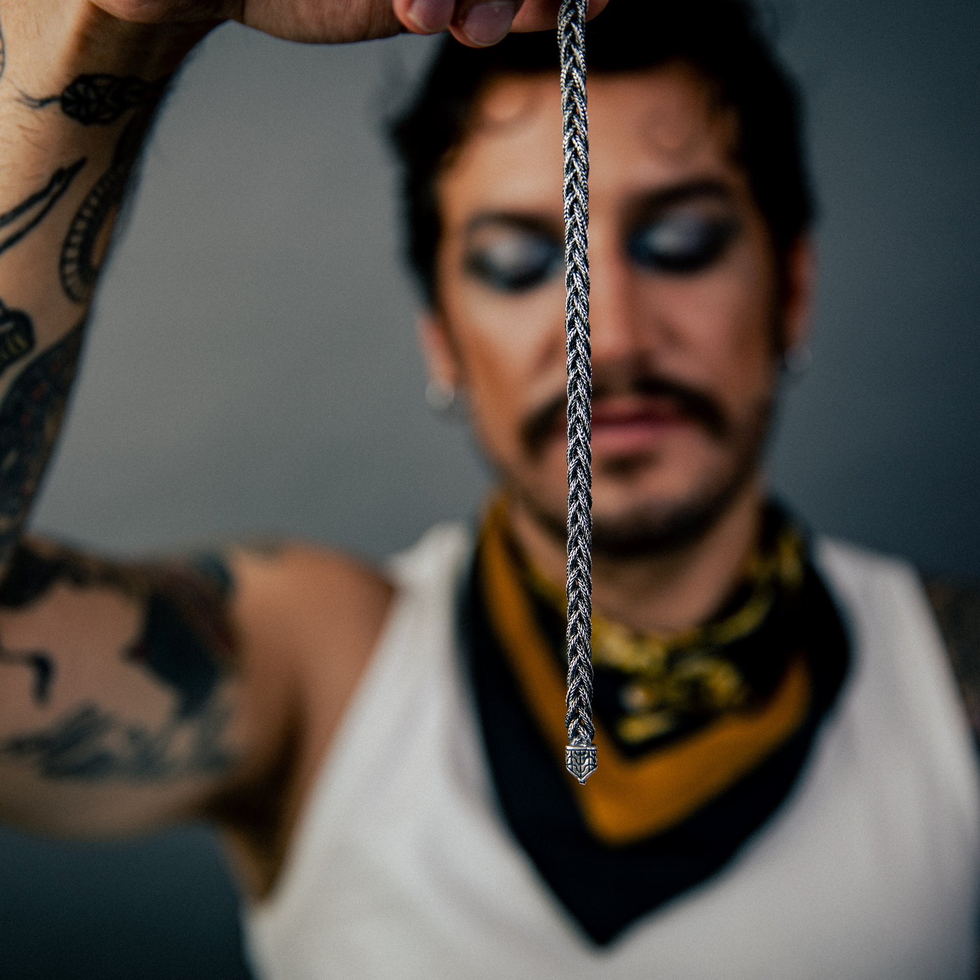 [full] - Person with tattoos holding a woven sterling silver bracelet with oxidised finish and intricate texture, in a blurred background