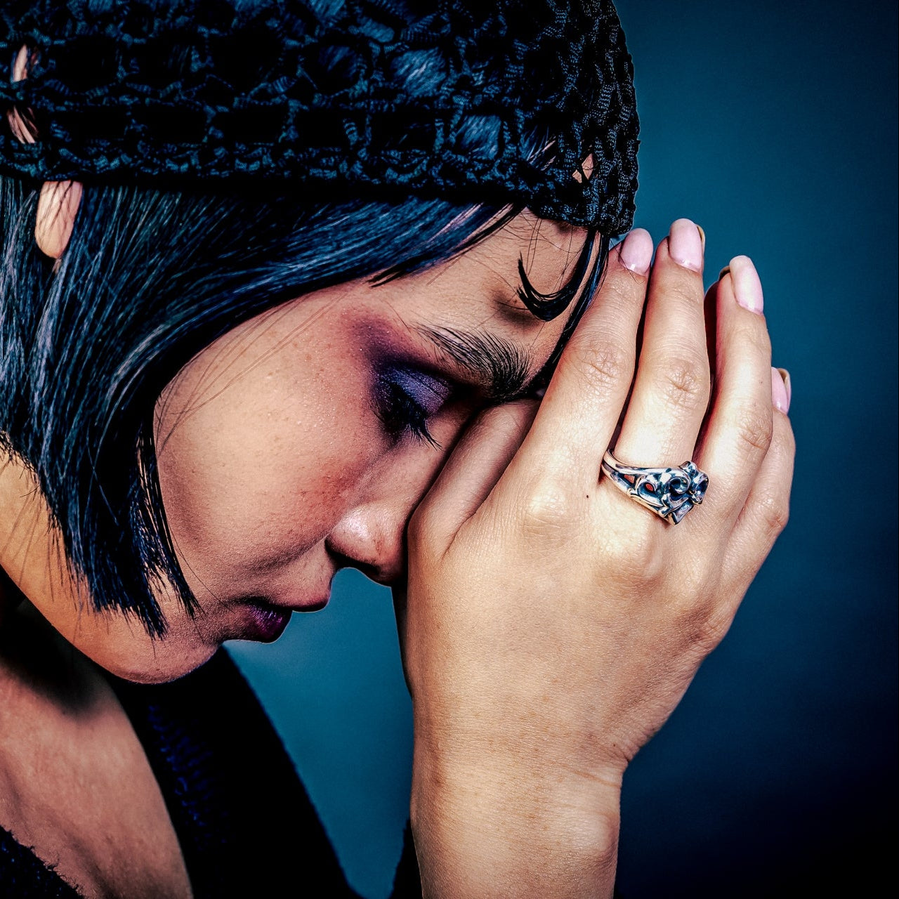 [full] Woman with short black hair wearing a black knit hat and black sweater, holding her head in her hands. A statement sterling silver ring is in one of her fingers